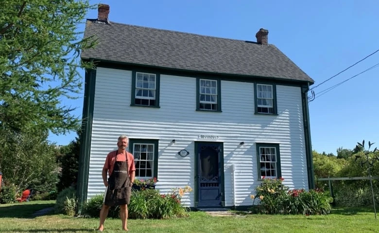 A man in an apron stands in front of a white two-story house with dark trim.