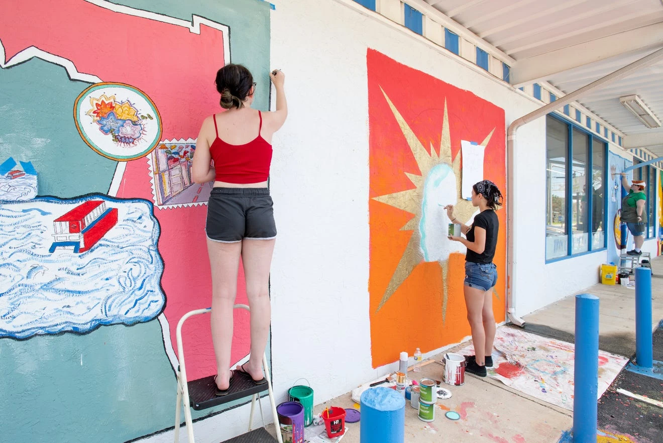 From left, University of West Florida students Kaylie Shultz, Whitney Hayek and Anna Ballow paint murals on the exterior of the Shoreline Foods International Market & Deli in Pensacola on Thursday.