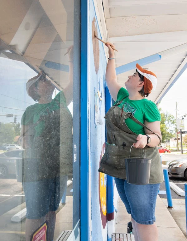 University of West Florida senior Anna Ballow paints a mural on the exterior of Shoreline Foods International Market & Deli in Pensacola on Thursday.